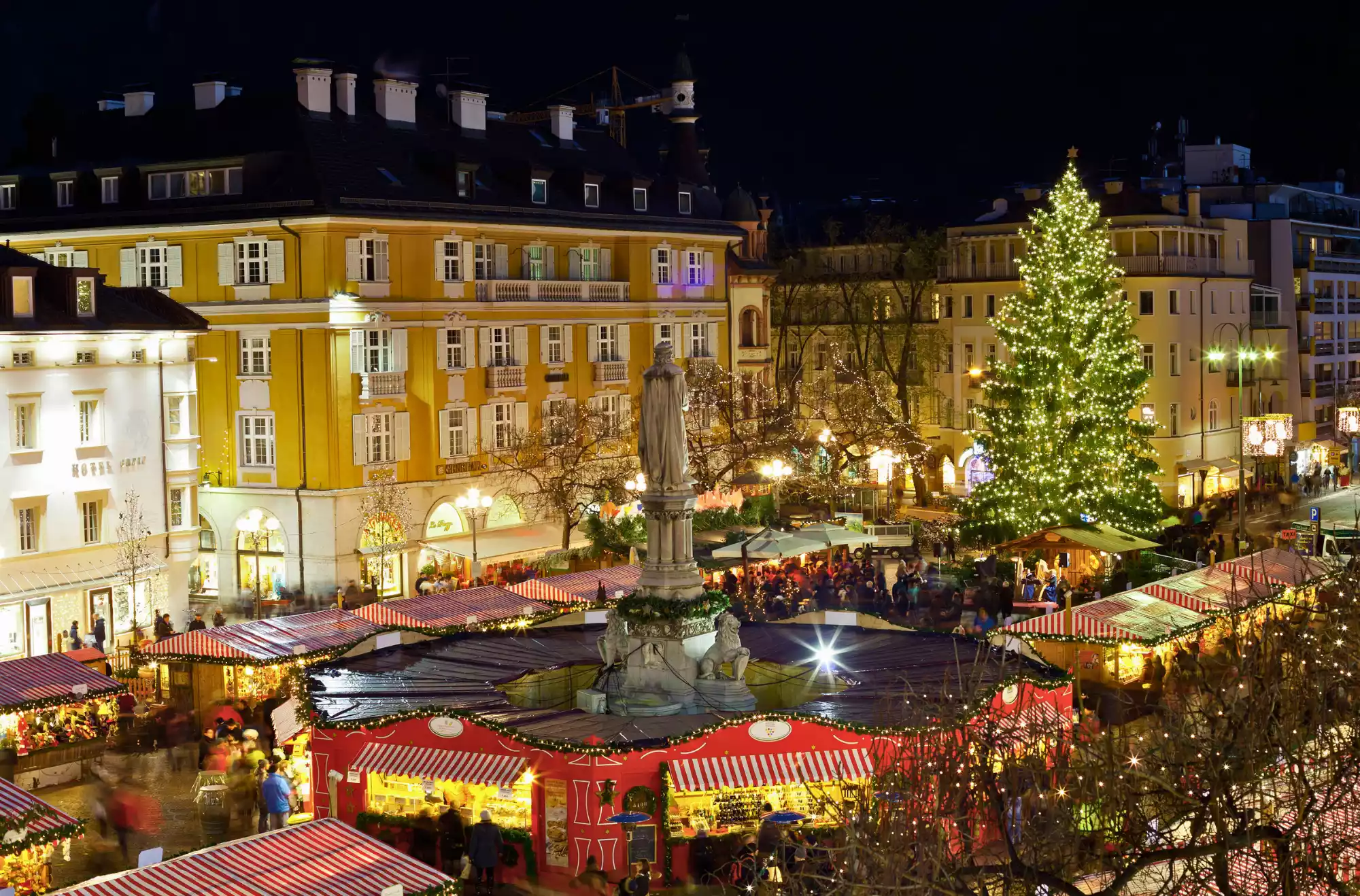 Veduta notturna del Mercatino di Natale in Piazza Walther a Bolzano. Un grande albero di Natale illuminato e le casette rosse e bianche circondano un monumento centrale, con i palazzi storici sullo sfondo