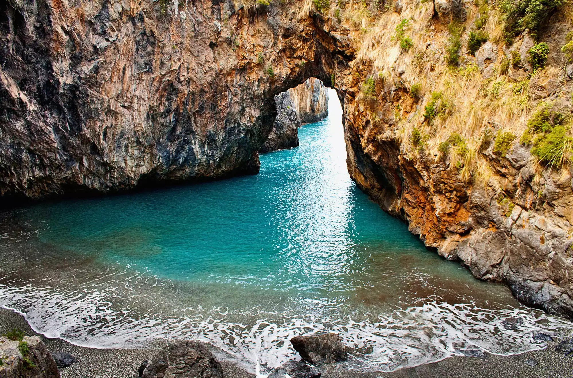 Veduta della suggestiva spiaggia dell’Arcomagno, incastonata tra alte pareti rocciose, con acqua cristallina e un arco naturale che si apre sul mare, lungo la costa tirrenica della Calabria.