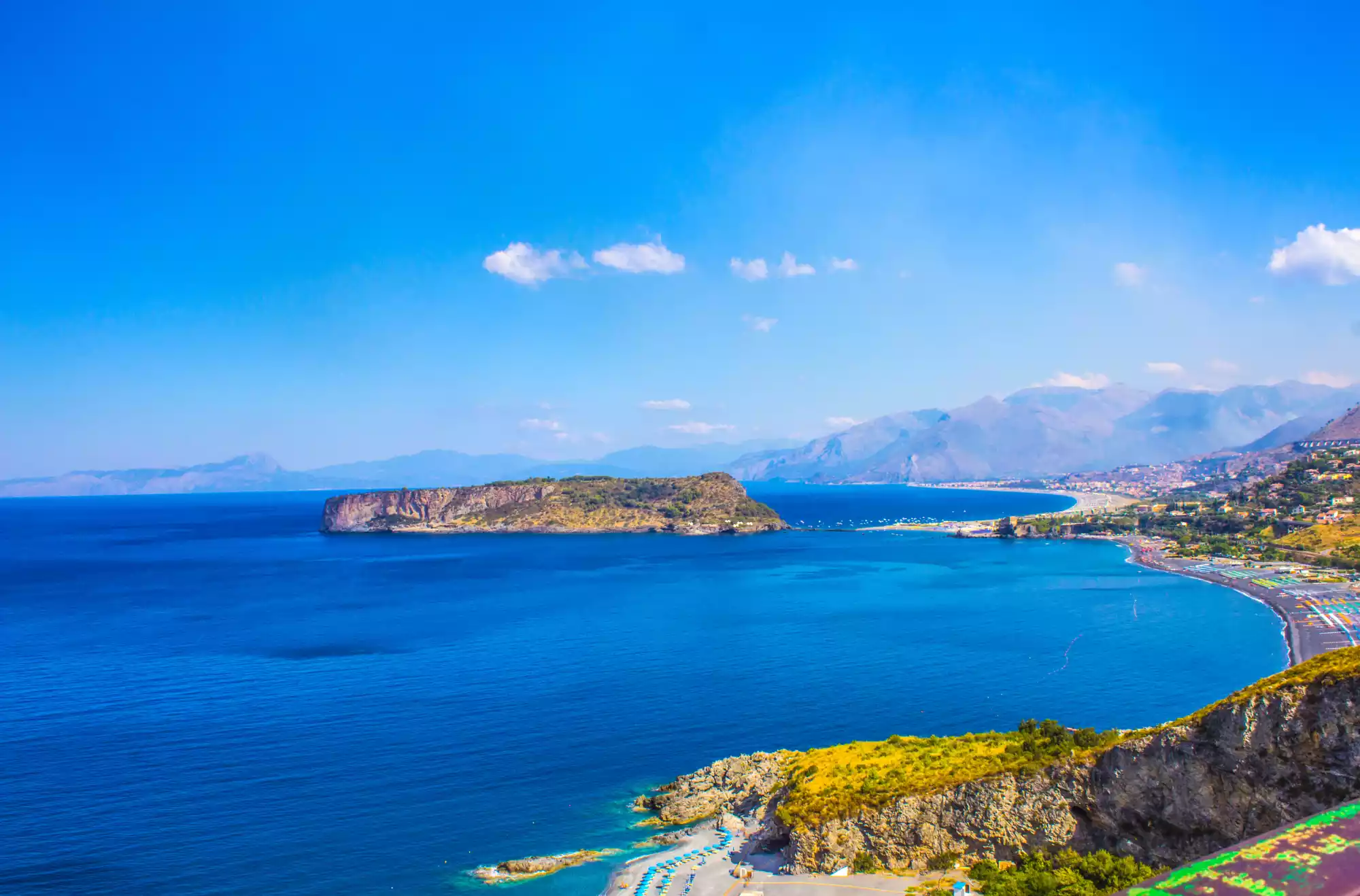 Vista panoramica sull’Isola di Dino e sul litorale calabrese, con acque cristalline, spiagge attrezzate e montagne sullo sfondo, in una splendida giornata di sole.