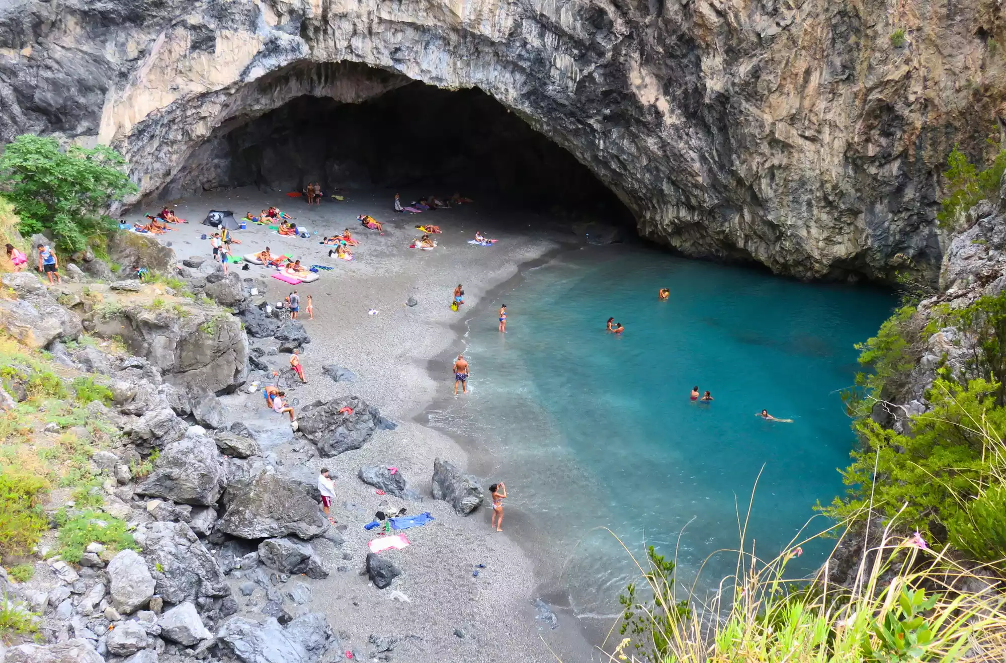 Grotta dell’Arcomagno a San Nicola Arcella affollata di bagnanti, con spiaggia di ciottoli e acque cristalline circondate da alte pareti rocciose.