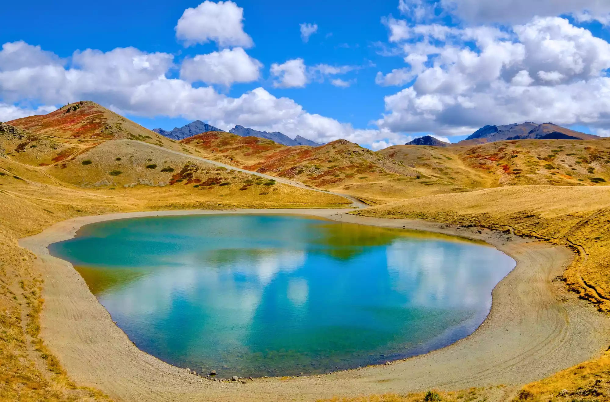 Foto del Lago di Gineaux in Val di Susa, Piemonte, un piccolo lago alpino dalle acque turchesi incorniciato da rilievi montuosi con praterie di montagna e un cielo azzurro con nuvole bianche.