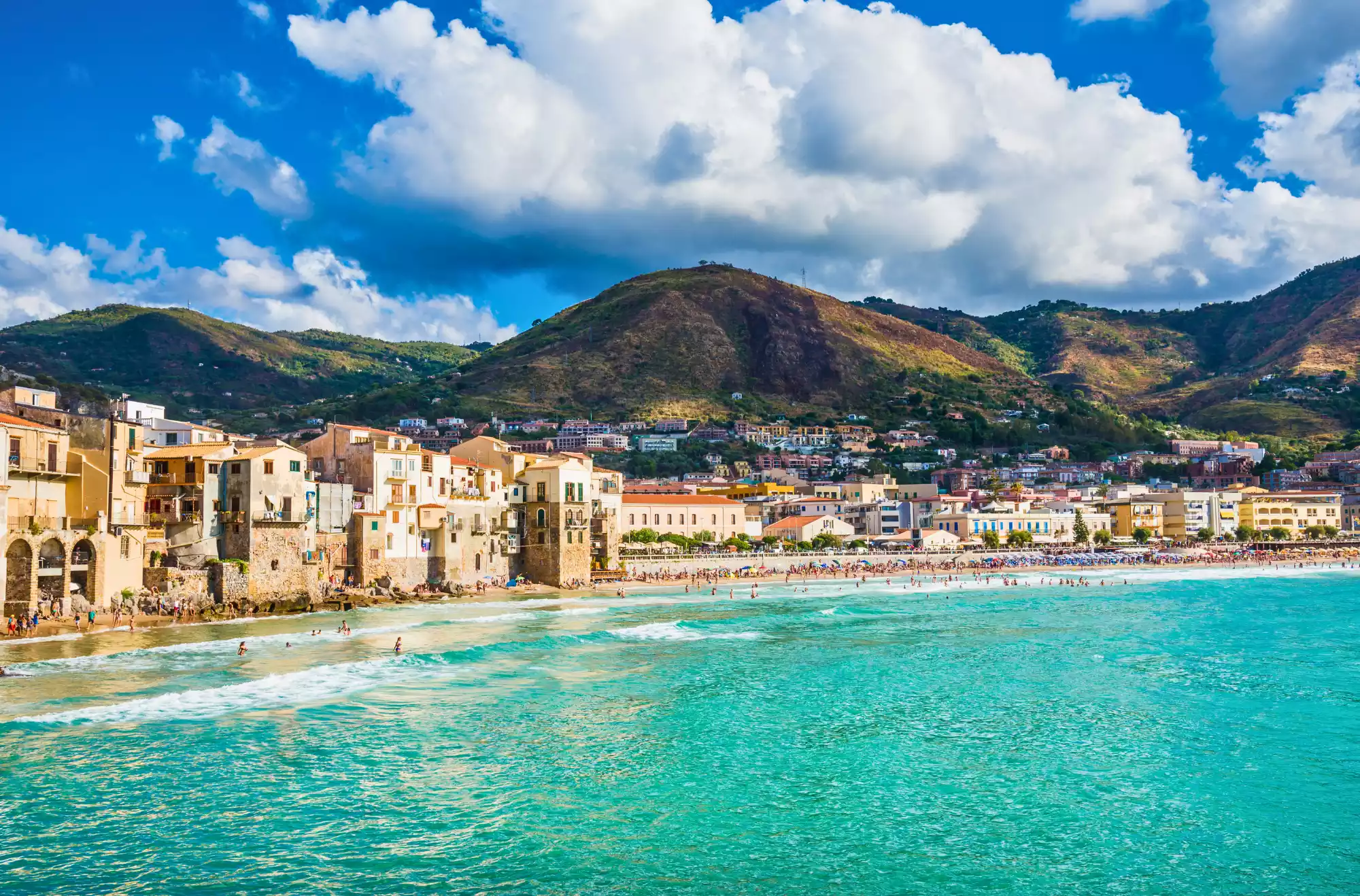 la splendida spiaggia di Cefalù in Sicilia con le sue acque cristalline e il litorale affollato. Sullo sfondo le colline verdi e un cielo azzurro tipico delle giornate estive.