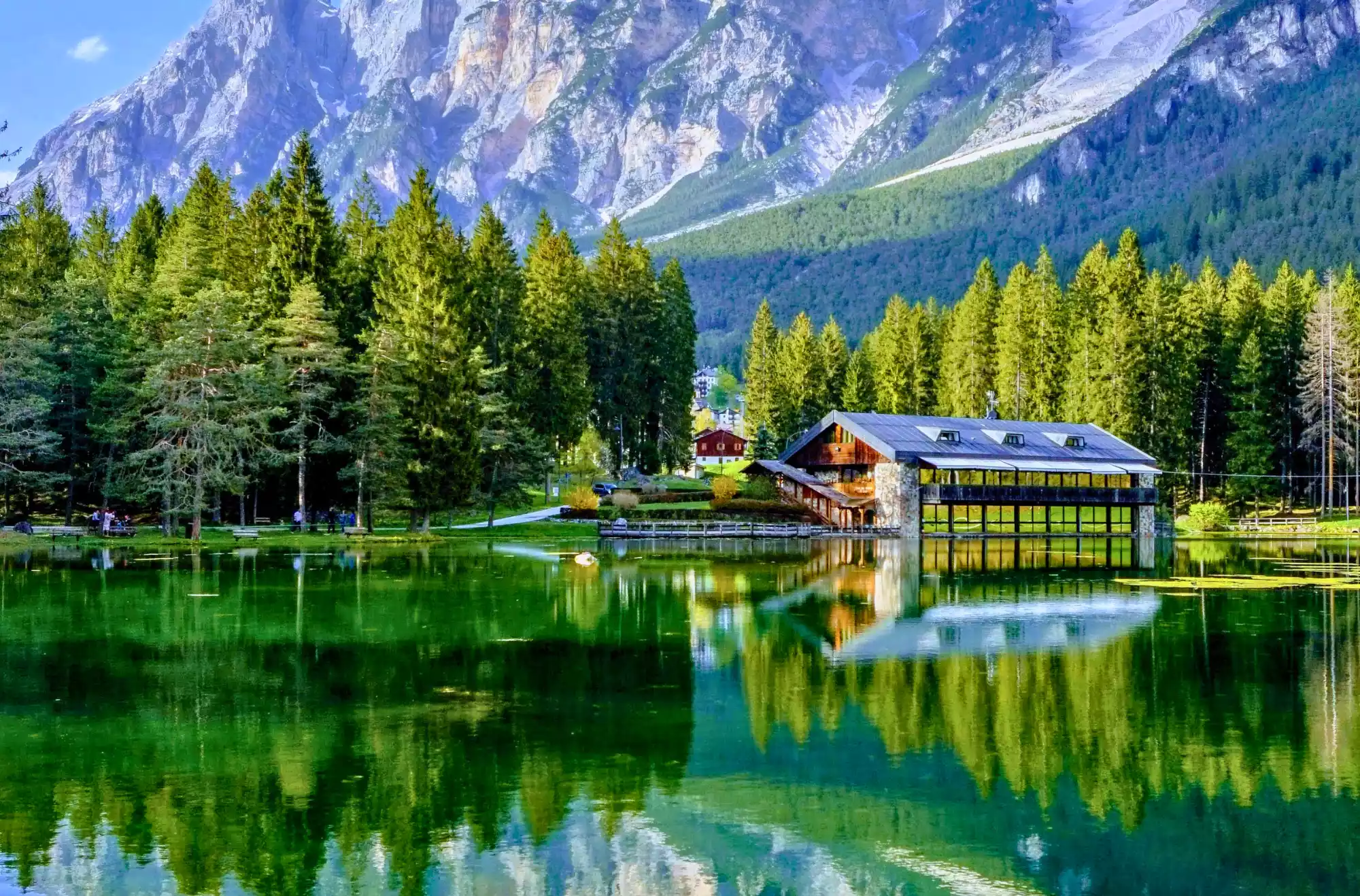 Riflessi verde smeraldo sull'acqua calma del Lago di Mosigo, in Veneto, che rispecchia un rifugio in legno con grandi finestre e gli abeti che lo circondano. Sulla riva opposta, si intravedono alcune case e la cima di una montagna rocciosa.