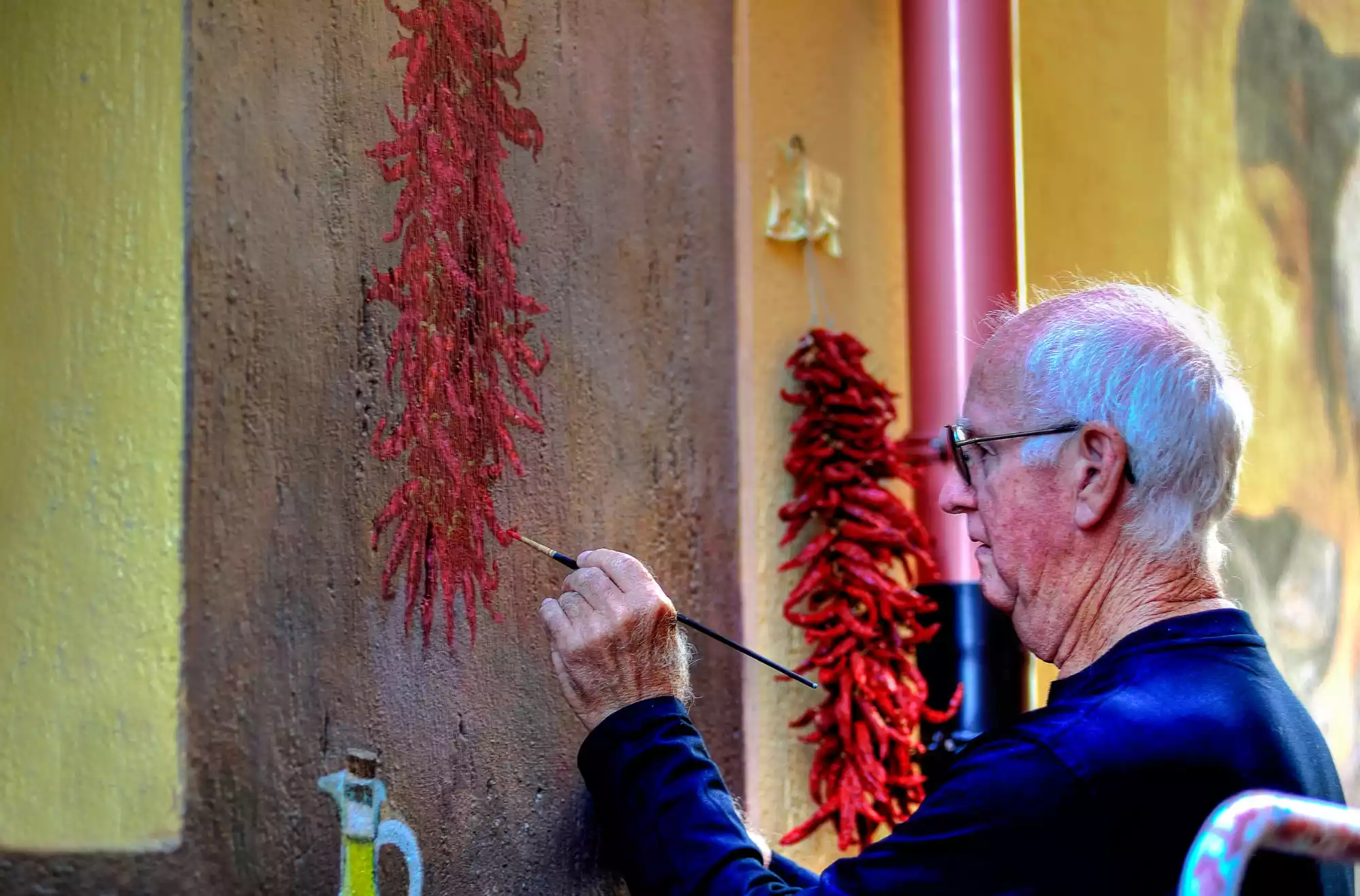 Uomo anziano dipinge un mazzo di peperoncini rossi su un muro, accanto a una parete con peperoncini veri appesi, in una scena che unisce arte e tradizione, tipica di Diamante, Calabria.