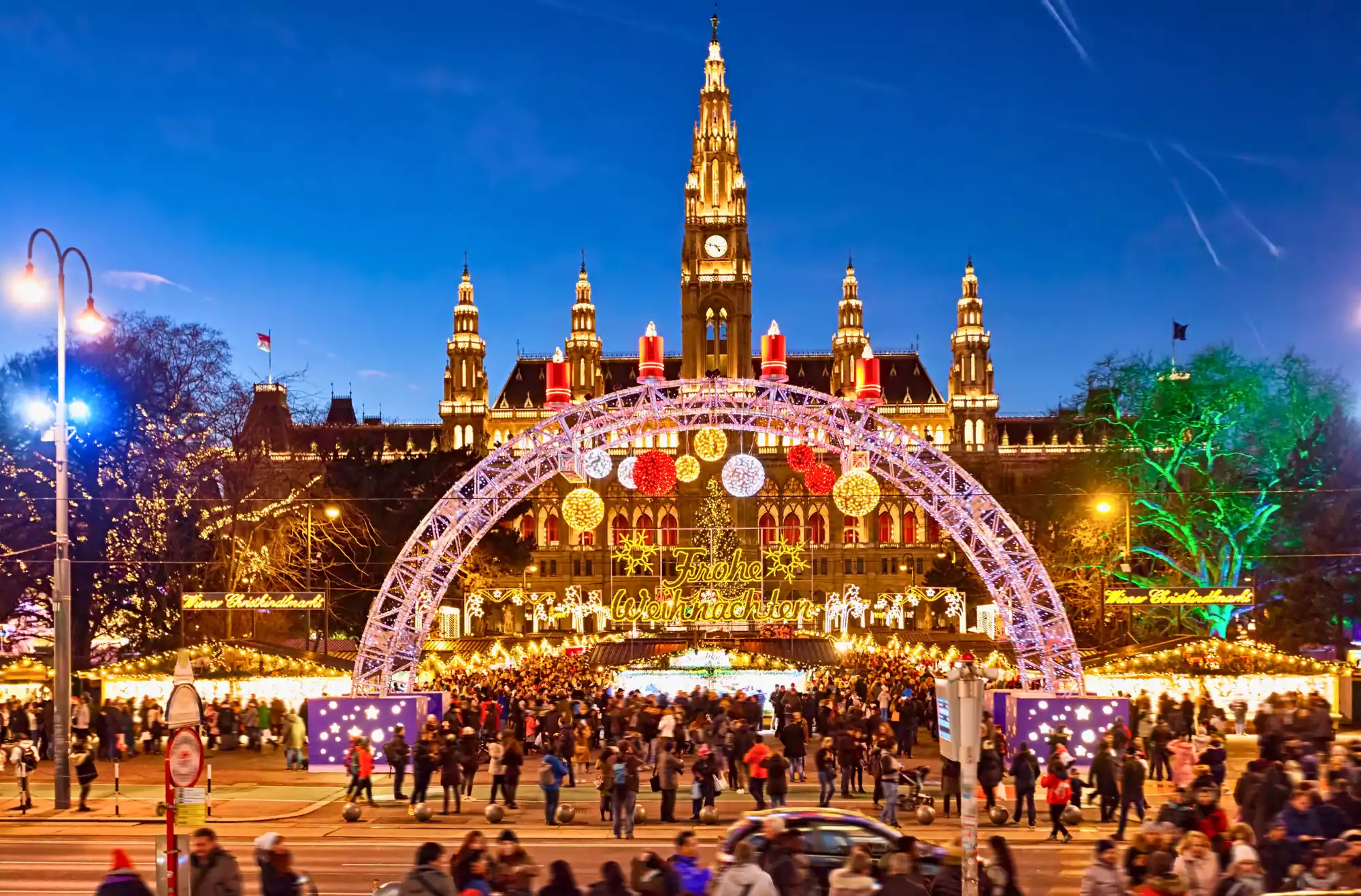 Vista notturna dei mercatini di Natale a Vienna di fronte al Rathaus, con una folla di persone e luminose decorazioni festive.
