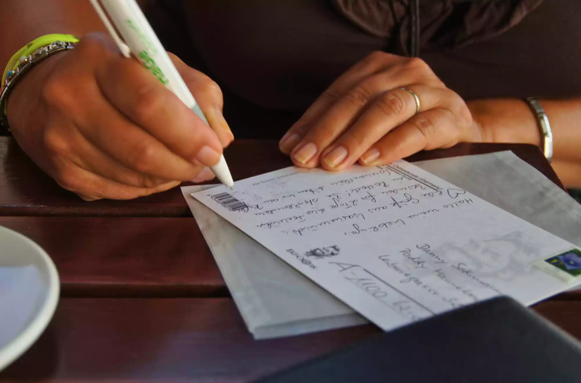 mani di una turista ripresa nell'atto di scrivere una cartolina di viaggio su un tavolo di legno