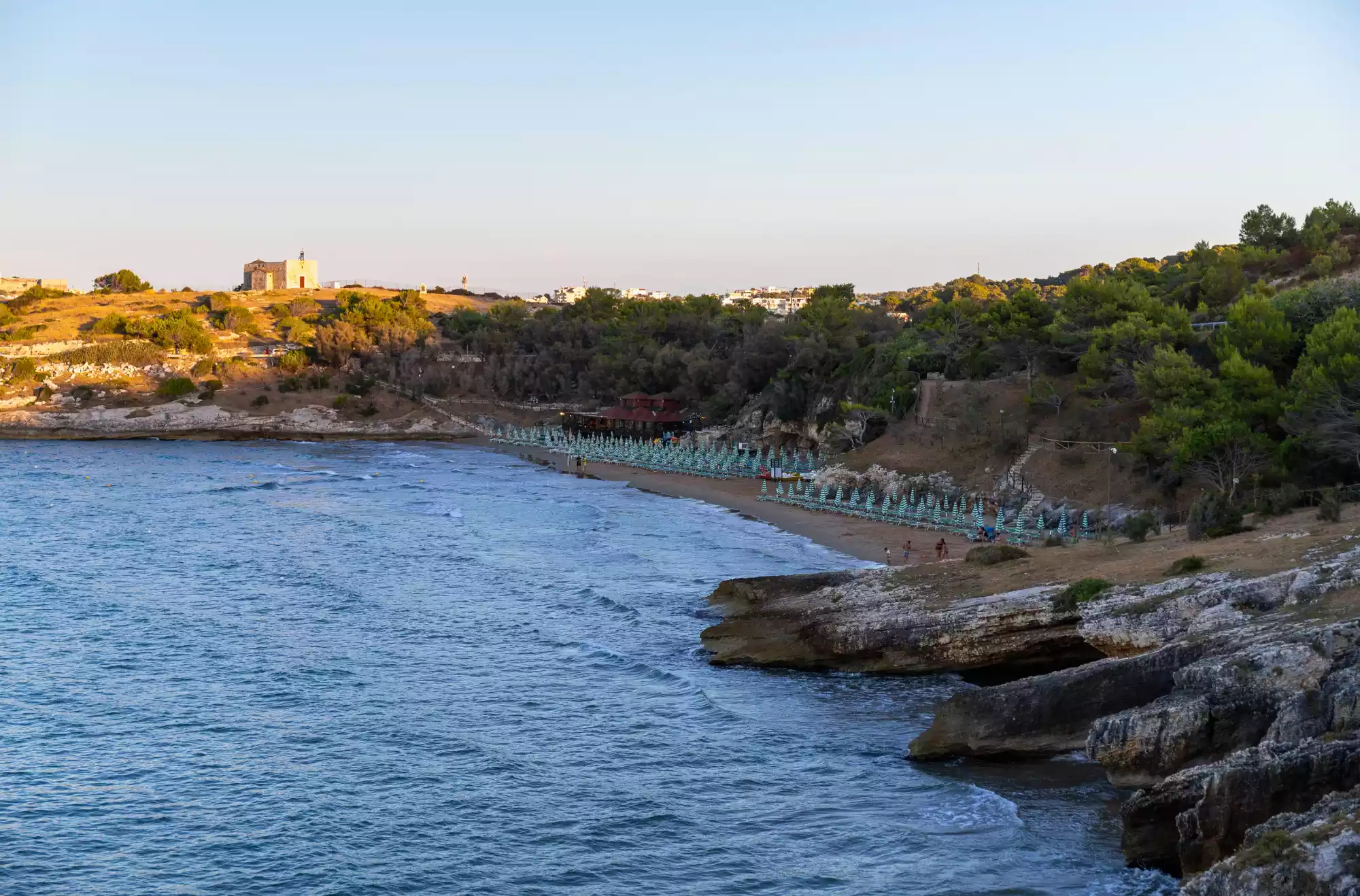 Spiaggia di San Lorenzo nel Gargano al tramonto, con ombrelloni allineati lungo la riva, mare calmo e scogliere rocciose in primo piano, circondata da vegetazione e colline.